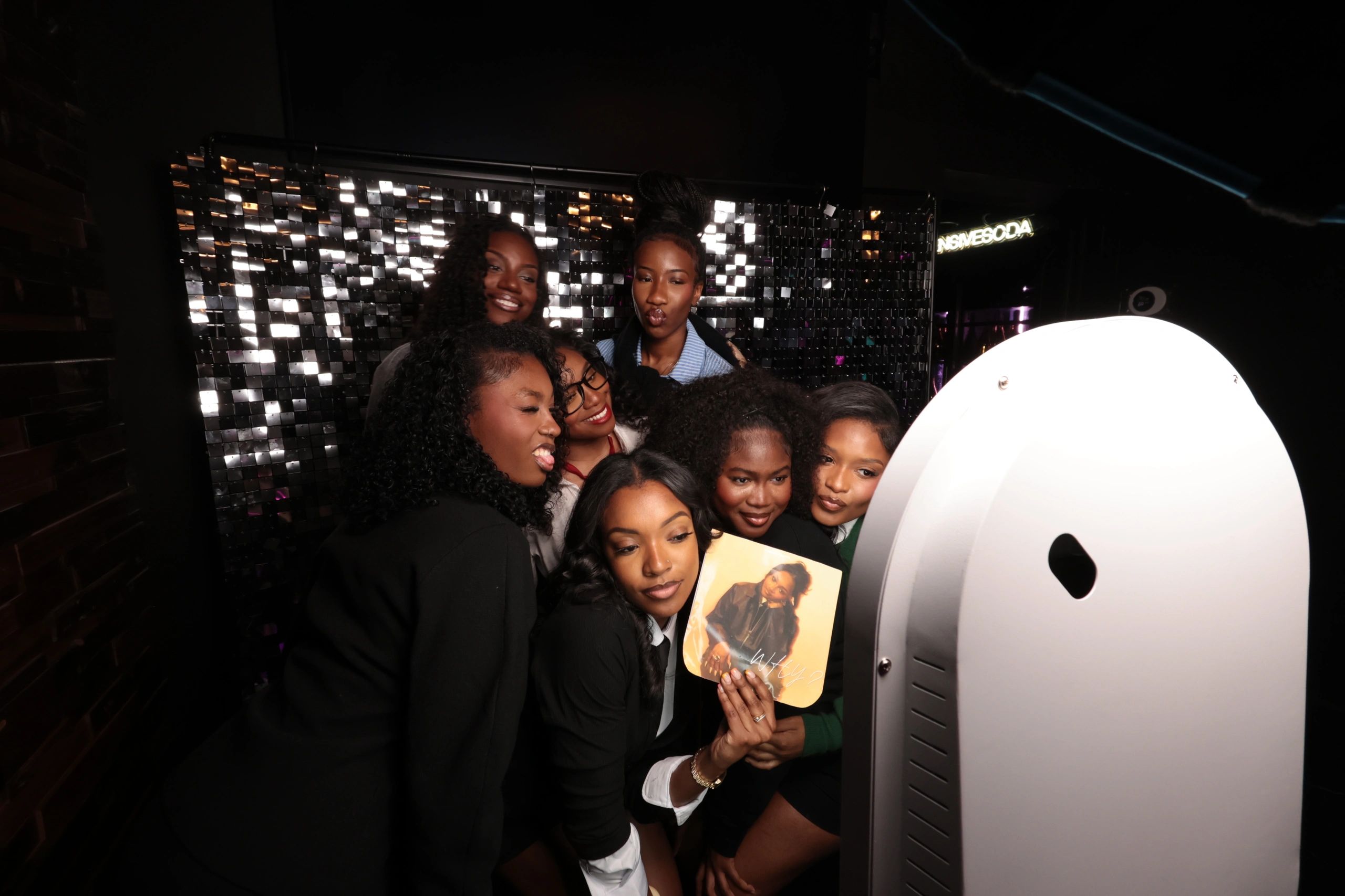 A group of friends joyfully taking a selfie in a photo booth with a sparkly backdrop.