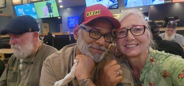 A smiling couple posing closely in a lively restaurant with TV screens in the background.