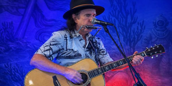 Musician playing guitar and harmonica on stage with vibrant blue and red lighting.