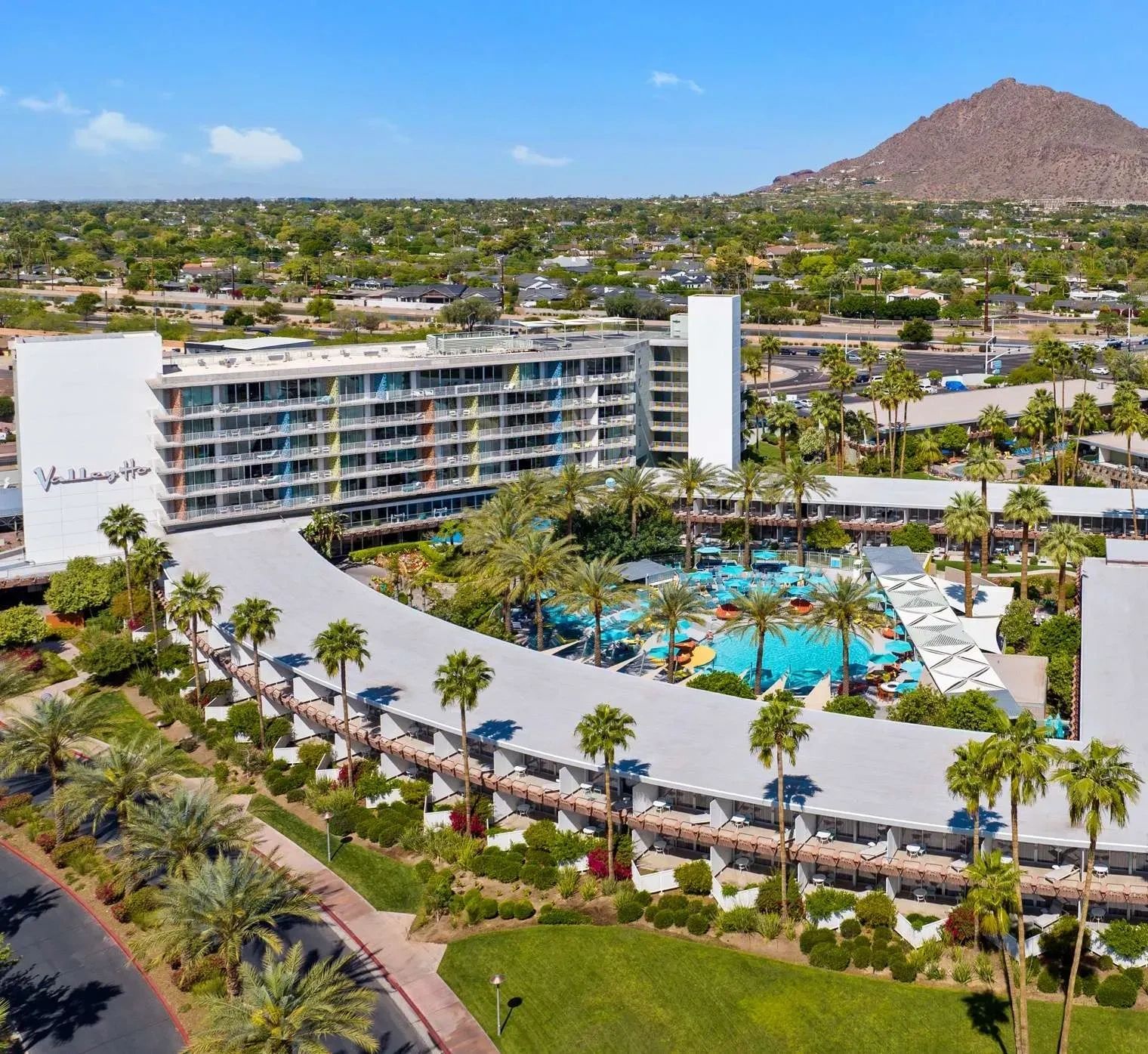 Aerial view of a curved hotel with a large pool and palm trees.