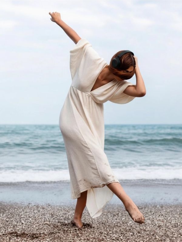 Woman dancing barefoot on a beach wearing a white dress and headphones.