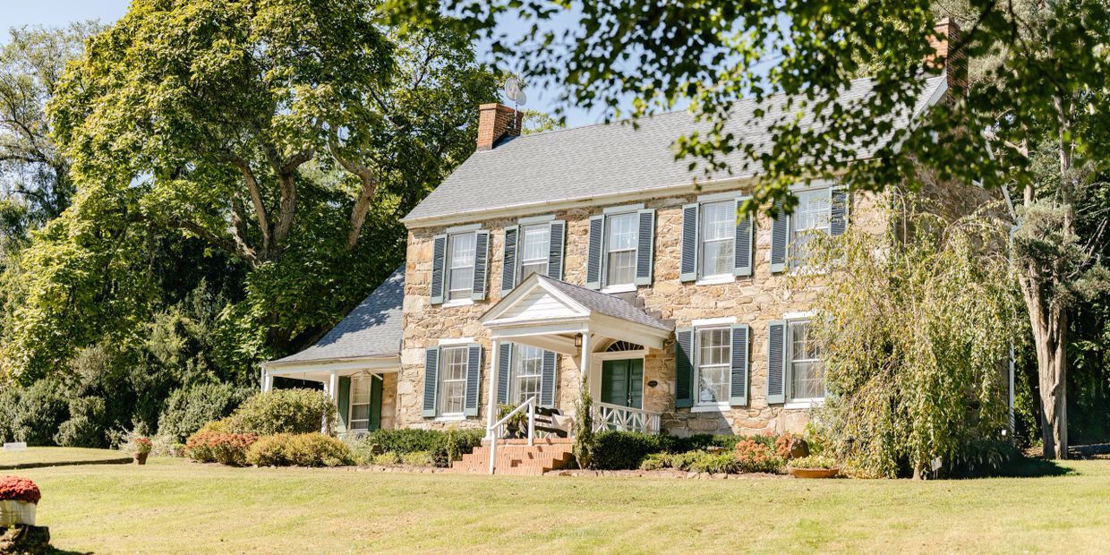 Stone house with green shutters surrounded by lush greenery and trees.
