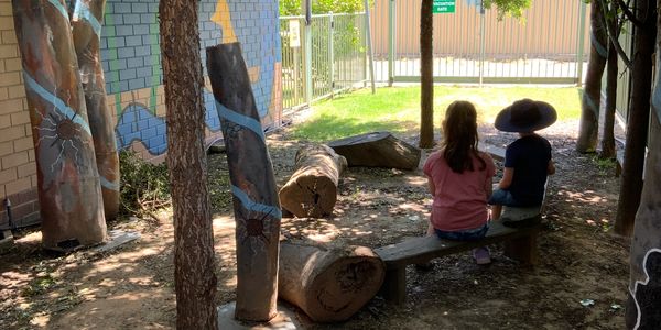 Two children sit on a bench in a shaded outdoor area with painted tree trunks.