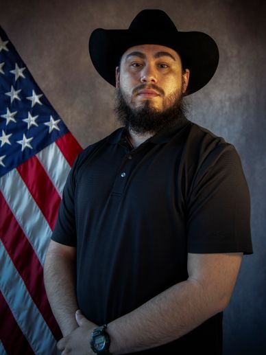 Man in black shirt and cowboy hat standing confidently by American flag.