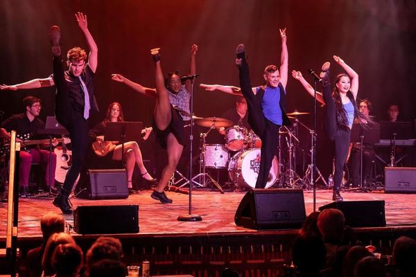 Four dancers performing high kicks on stage with a live band behind them.