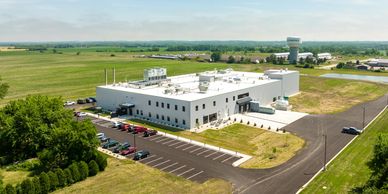 A modern industrial building surrounded by green fields and parking lots.