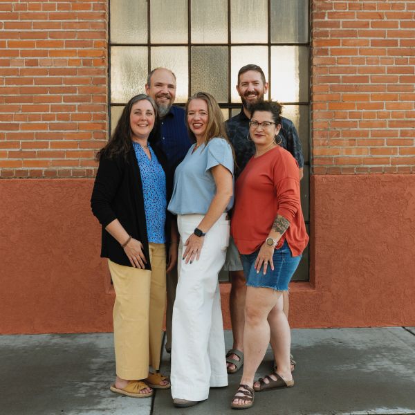 Group of five adults posing happily in front of a brick wall.