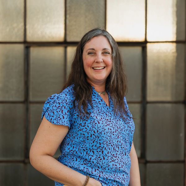 Smiling woman in a blue patterned blouse, standing in front of a window.