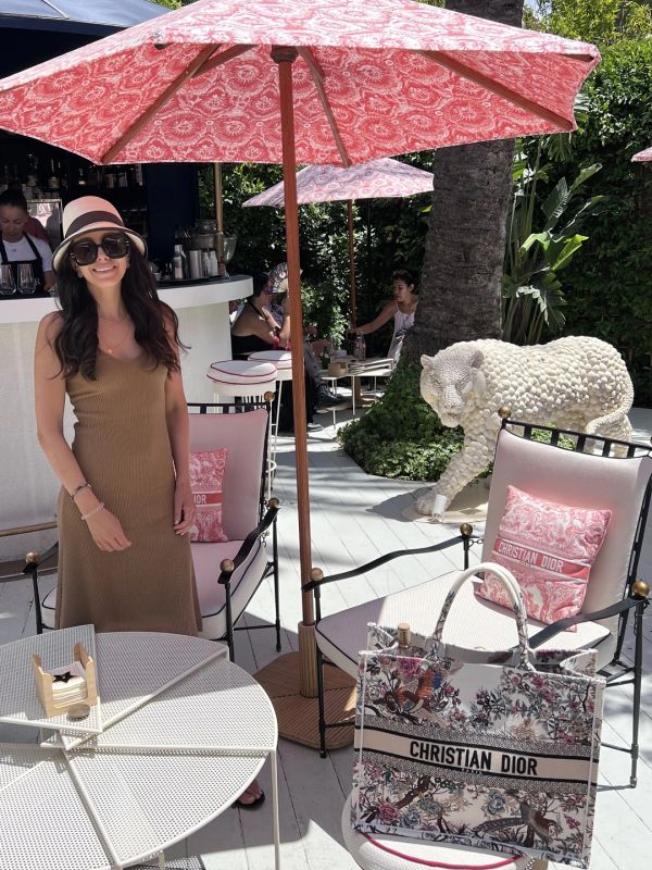 Woman in a hat standing under a pink umbrella at a chic outdoor cafe.