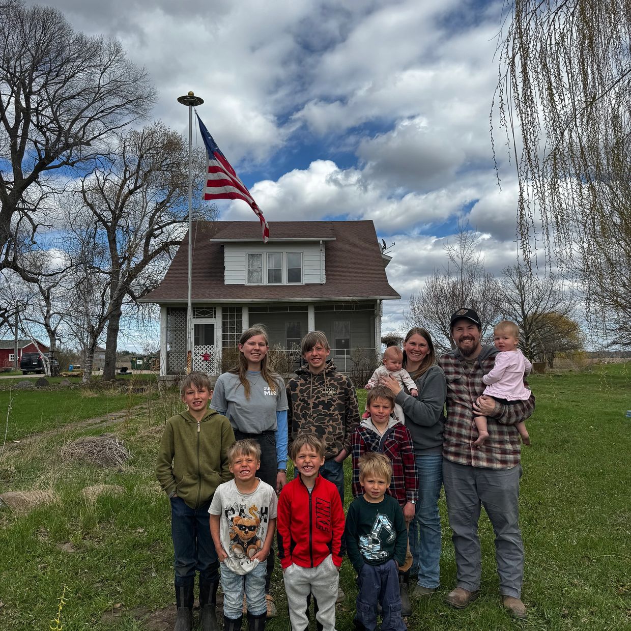 A large family posing in front of their house with an American flag on a cloudy day.