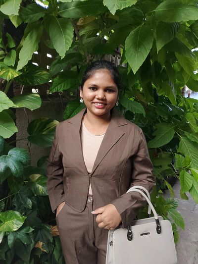 A smiling woman in a brown suit stands in front of lush green leaves.