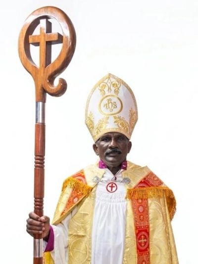 A bishop in ornate religious vestments holding a wooden crozier with a cross.