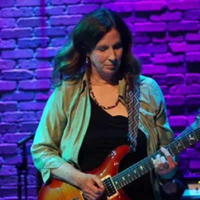Woman playing electric guitar on stage with purple lighting.