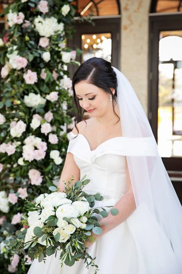 Bride in white dress holding bouquet, standing by floral arch.