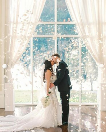 Bride and groom kissing indoors with flower petals falling.