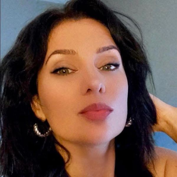 A Romanian-American woman purses her lips. Wearing twisted, gold hoops and heavy makeup. 