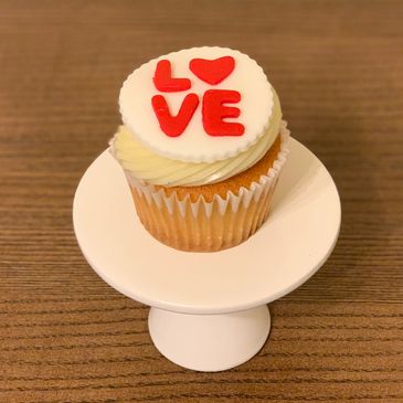 Cupcake with white frosting and red love decoration on a white stand.