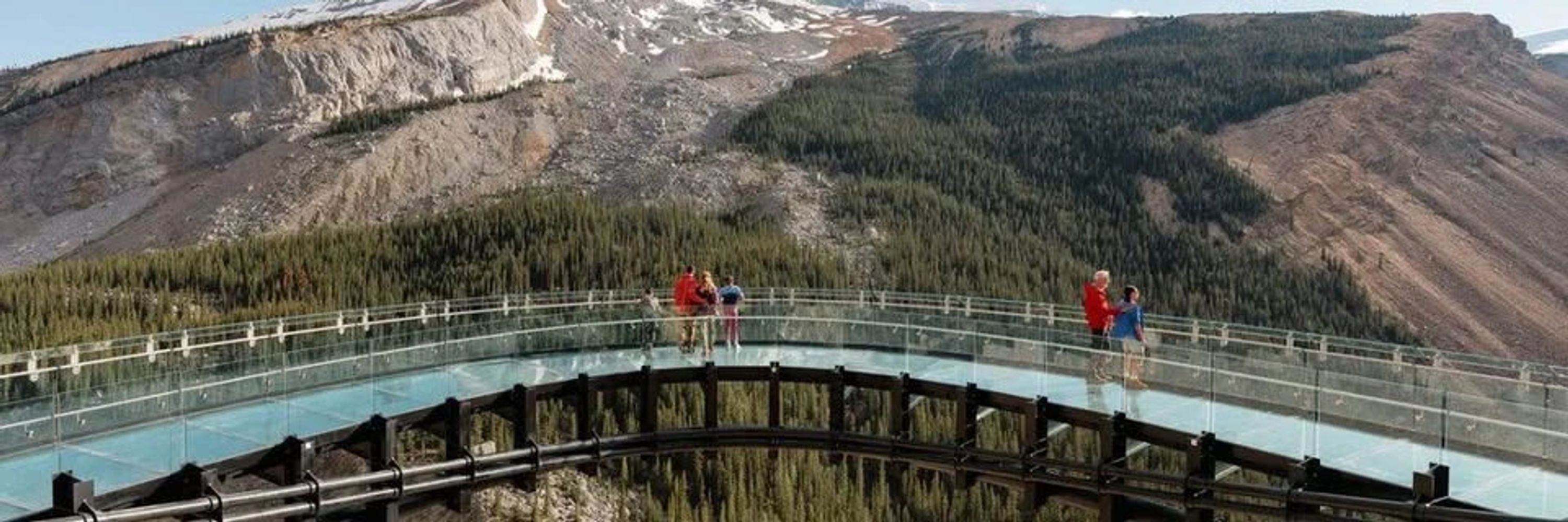 Curved glass bridge with people overlooking mountainous forest scenery.