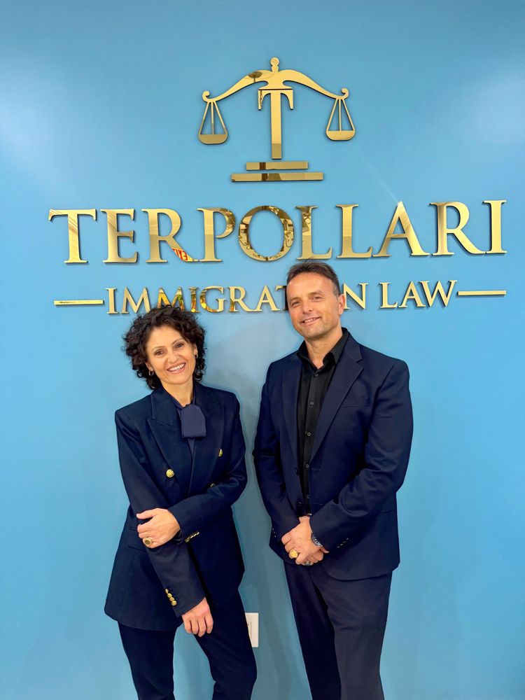 Two professionals posing in front of a blue wall with a gold immigration law sign.