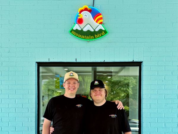 Two men in black shirts and hats stand smiling under a colorful chicken logo on a blue wall.