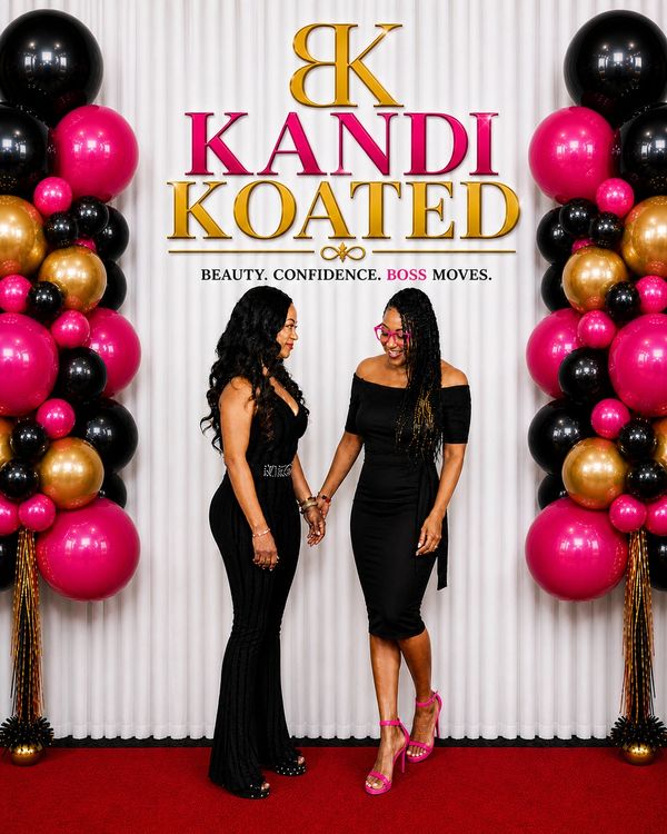 Two elegantly dressed women holding hands at a Kandi Koated event with bold balloon decor.