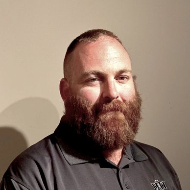 Bearded man with a shaved head in a black polo shirt against a plain wall.
