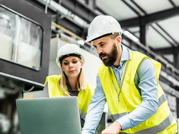 Two engineers in safety gear reviewing plans on a laptop in a factory.