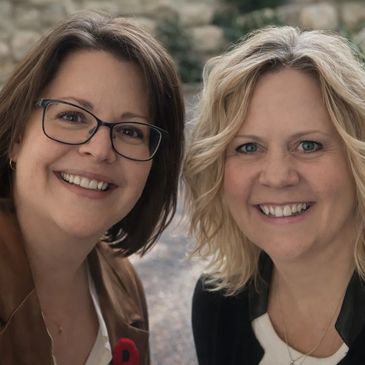 Two women smiling warmly outdoors, one wearing glasses and brown jacket.