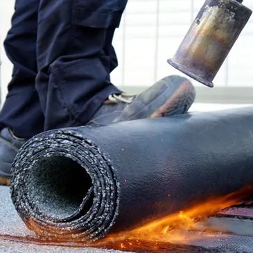 Worker using a torch to install rolled roofing material on a flat roof.