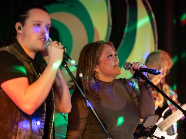Two singers performing on stage with colorful lights and a guitarist in the background.