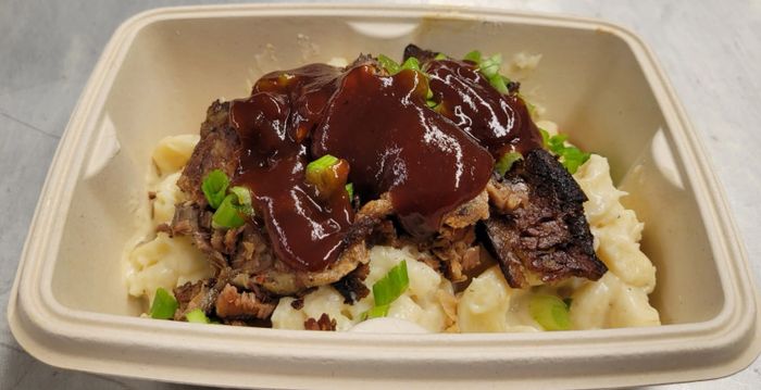 Barbecue brisket topped with sauce and green onions over creamy mac and cheese in a takeout container.