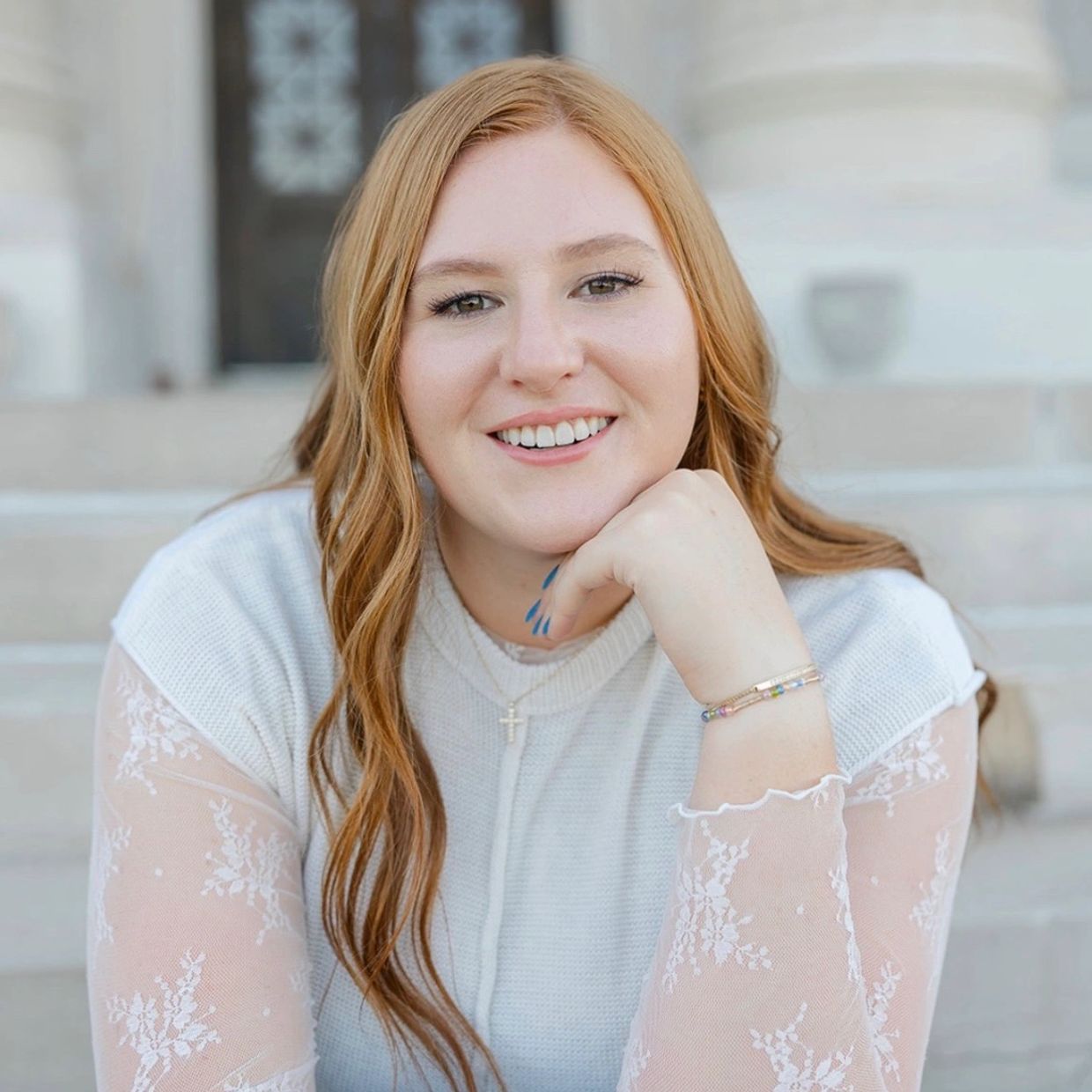 Smiling young woman with red hair and a lace-sleeved top poses outdoors.