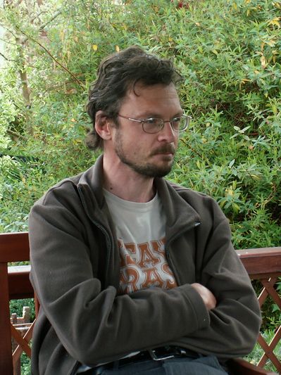 Man with glasses sitting outside on a wooden bench with arms crossed.