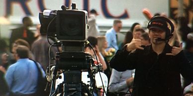 A cameraman gestures while filming in a crowded outdoor setting.