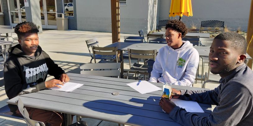 Three young men study outdoors at a table on a sunny day.