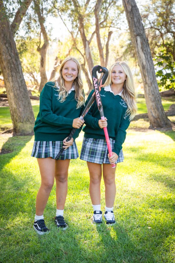 Two girls in school uniforms holding field hockey sticks outdoors.
