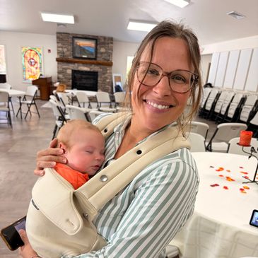 A smiling woman holding a sleeping baby in a carrier inside a community hall.