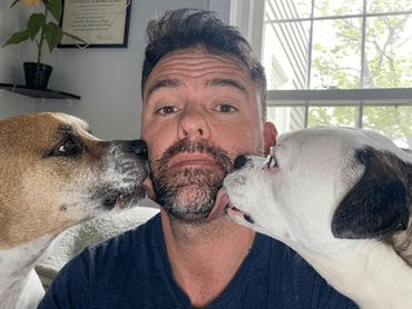 Roy sitting with his 2 dogs on each side of him licking his face as he looks straight ahead. 