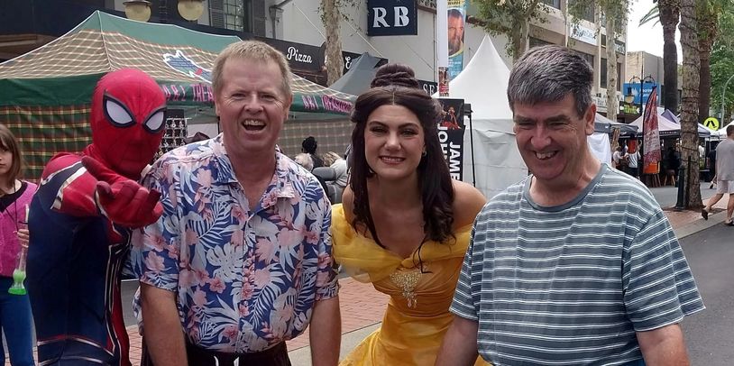People posing with Spider-Man and a woman in a yellow princess dress at an outdoor event.