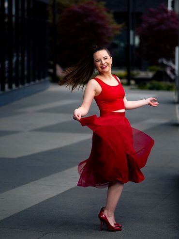 A white woman in bright red dances in the street of a city. Her tulle skirt and long dark ponytail s