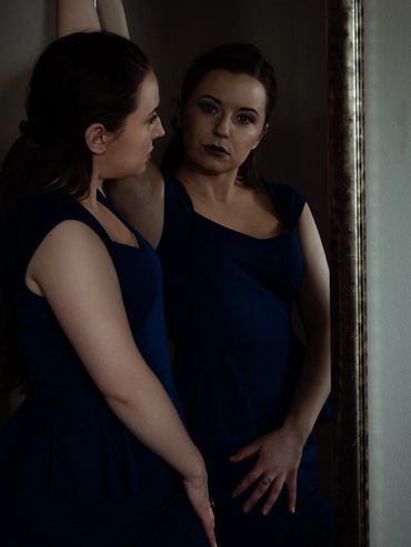 A white woman with long dark hair looks into a mirror at the camera. She rests her hand on her hip a