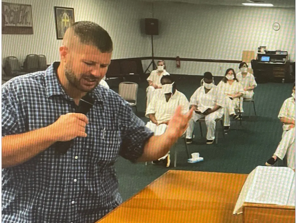Man passionately speaking into a microphone in front of seated people in white uniforms.