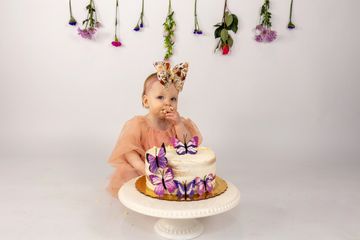 A baby girl with a floral headband sitting behind a butterfly-themed cake.