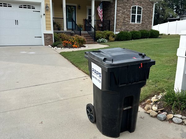 A black trash bin on a driveway near a house.