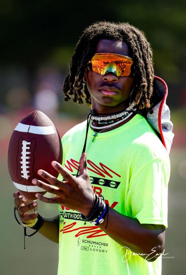 Young athlete in bright shirt holding a football, wearing reflective sunglasses and necklaces.