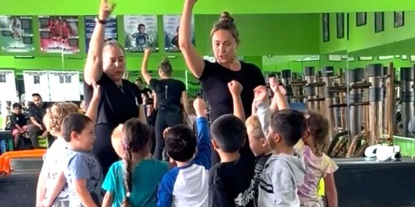 Two instructors lead a group of children in a gym activity, raising their hands together.