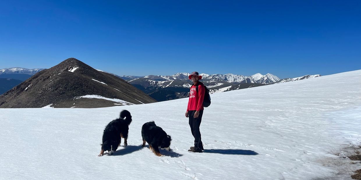 Man in red with two dogs on snowy mountain under clear blue sky.