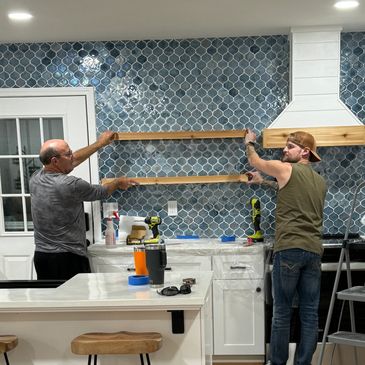 Two men installing wooden shelves on a blue tiled kitchen wall.