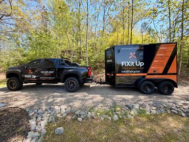 A black truck towing a branded trailer for FIXit Up in a wooded area.