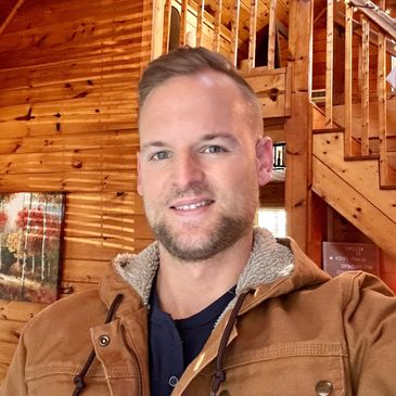 Man smiling inside a cozy wooden cabin with a staircase in the background.